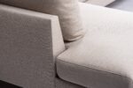 Close-up van een moderne beige stoffen bank met de armleuning, het rugkussen en het zitkussen. De stof heeft een subtiele textuur en het ontwerp is strak met strakke lijnen, wat het vakmanschap van de bank benadrukt.