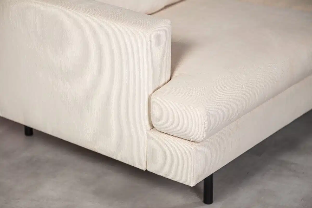 Close-up van een beige corduroy stoffen bank met een modern design, met een opvallende armleuning en zwarte metalen poten op een lichtgrijze vloer.