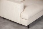 Close-up van een beige corduroy stoffen bank met een modern design, met een opvallende armleuning en zwarte metalen poten op een lichtgrijze vloer.