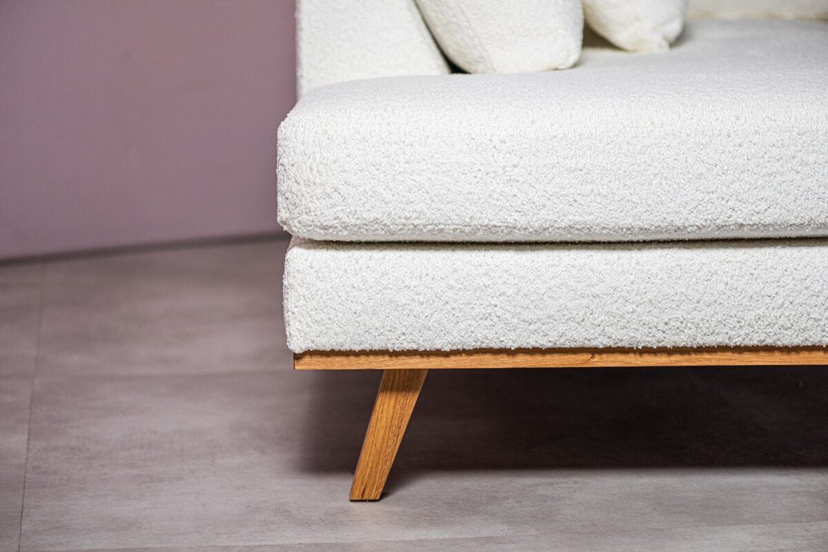 Close-up van een moderne witte getextureerde hoekbank met een houten poot op een lichtgrijze vloer. Een klein deel van een kussen is zichtbaar op de zitting, wat bijdraagt aan de gezellige en eigentijdse uitstraling. De achtergrondmuur is een gedempte tint roze.