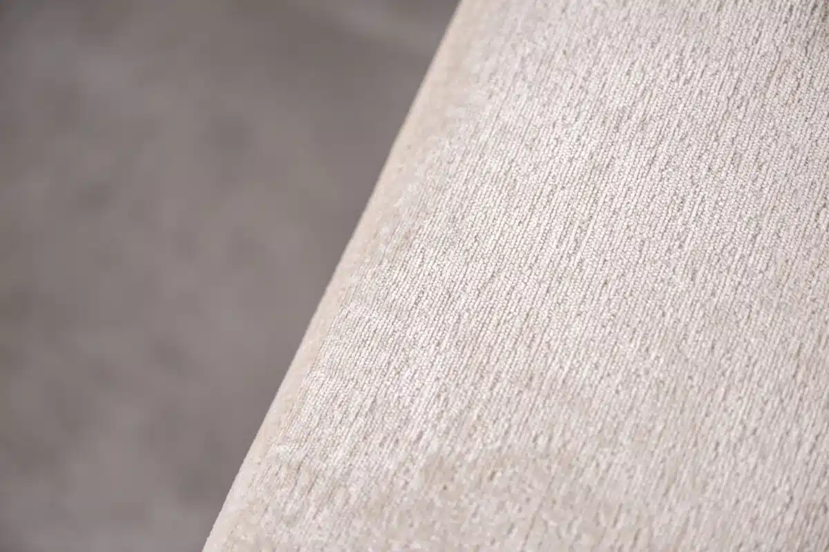 Close-up van een beige stof met een subtiel gestructureerd patroon. Het oppervlak lijkt zacht en loopt schuin van rechtsboven naar linksonder, waardoor het grootste deel van het frame wordt opgevuld.