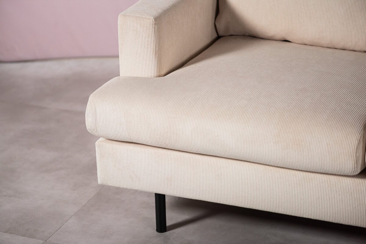 Close-up van een beige corduroy bank met een modern design, met een rechthoekige armleuning en een enkele zwarte metalen poot op een lichtgekleurde tegelvloer. De achtergrond is zacht vervaagd.