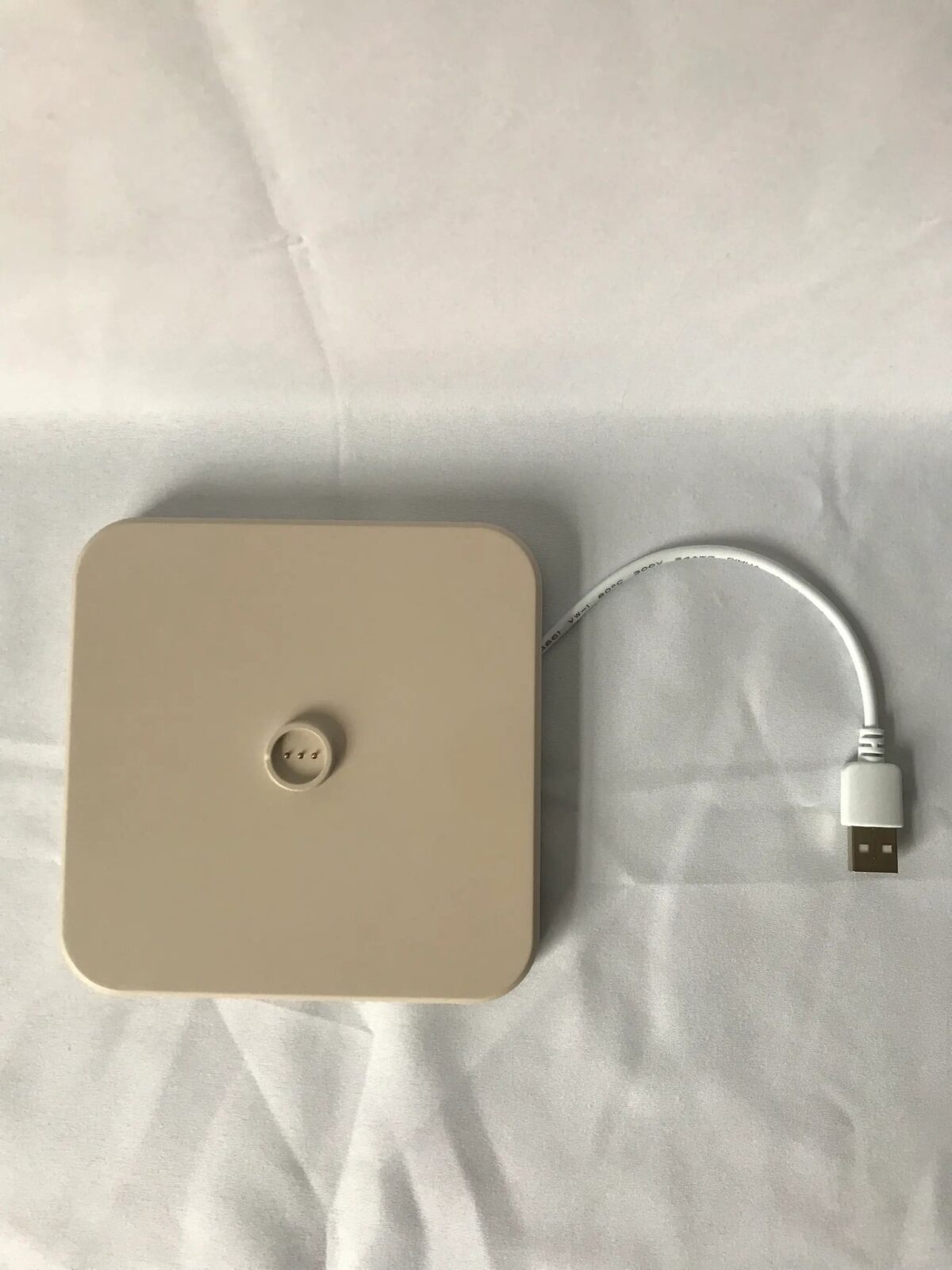 Een beige, vierkant elektronisch apparaat met een centrale knop staat op een wit oppervlak. Een witte kabel loopt vanaf de rechterkant en eindigt in een USB-connector.