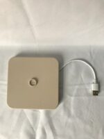 Een beige, vierkant elektronisch apparaat met een centrale knop staat op een wit oppervlak. Een witte kabel loopt vanaf de rechterkant en eindigt in een USB-connector.