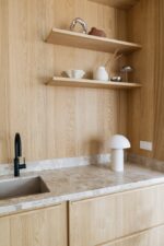 Minimalistische keuken met lichte houten kasten en marmeren aanrechtblad. Met een zwarte kraan, witte lamp en houten planken met decoratieve items zoals een theepot, vazen en rietverspreider tegen een met hout beklede muur.