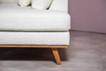 Close-up van een witte getextureerde bank met houten poten, geplaatst tegen een lichtpaarse muur. De vloer is betegeld in lichtgrijs. De afbeelding richt zich op de hoek van de bank en de moderne designelementen.