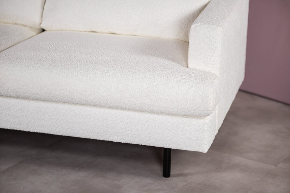 Close-up van een witte, getextureerde bank met een modern design, met een rechterarmleuning en een zwarte poot. De bank staat tegen een neutrale achtergrond met een beige vloer en een lichtpaarse muur.