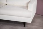 Close-up van een witte, getextureerde bank met een modern design, met een rechterarmleuning en een zwarte poot. De bank staat tegen een neutrale achtergrond met een beige vloer en een lichtpaarse muur.