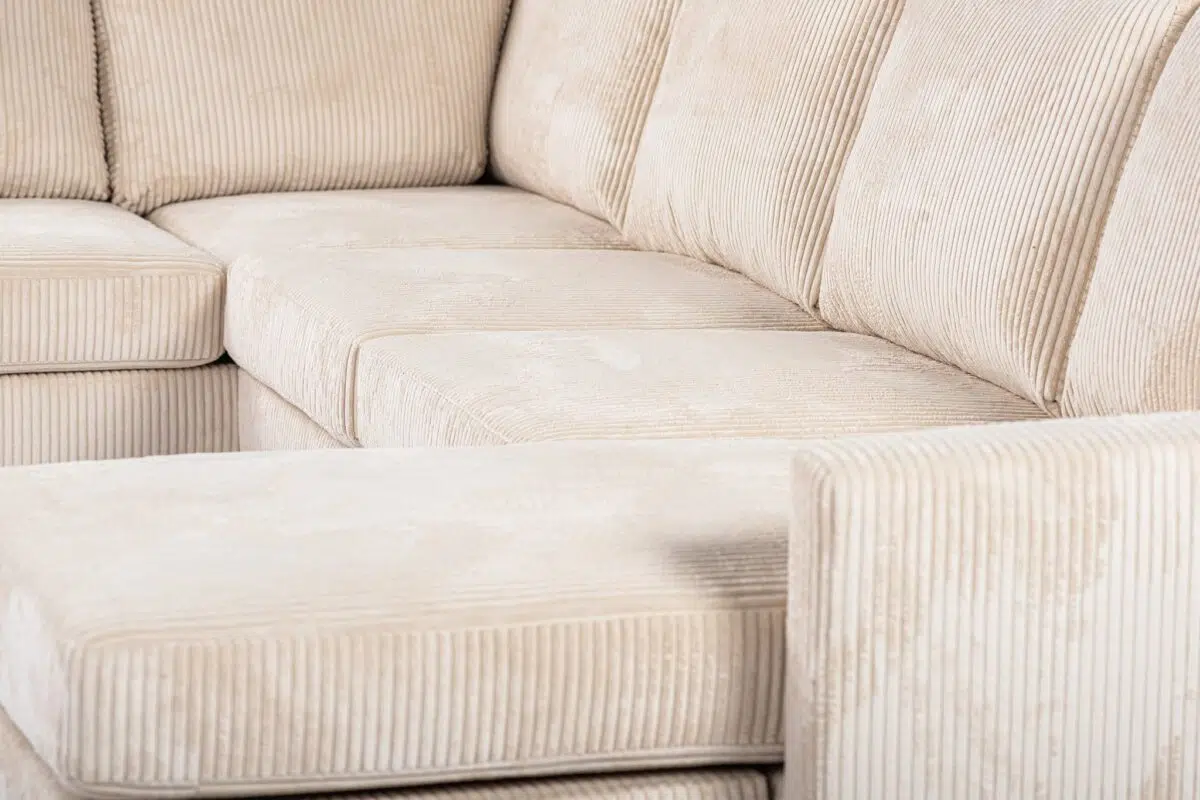 Close-up van een beige corduroy sectionele bank met zichtbare textuur en patroon. De bank heeft een hoekontwerp met diepe kussens en rechte lijnen, wat een moderne en gezellige uitstraling benadrukt.