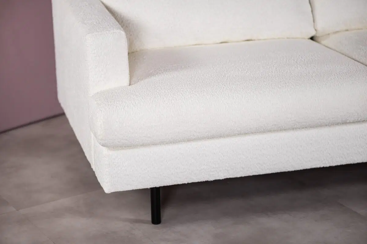 Close-up van een witte, getextureerde bank met strakke lijnen en zwarte poten, tegen een neutrale tegelvloer en een lichtgekleurde muur.