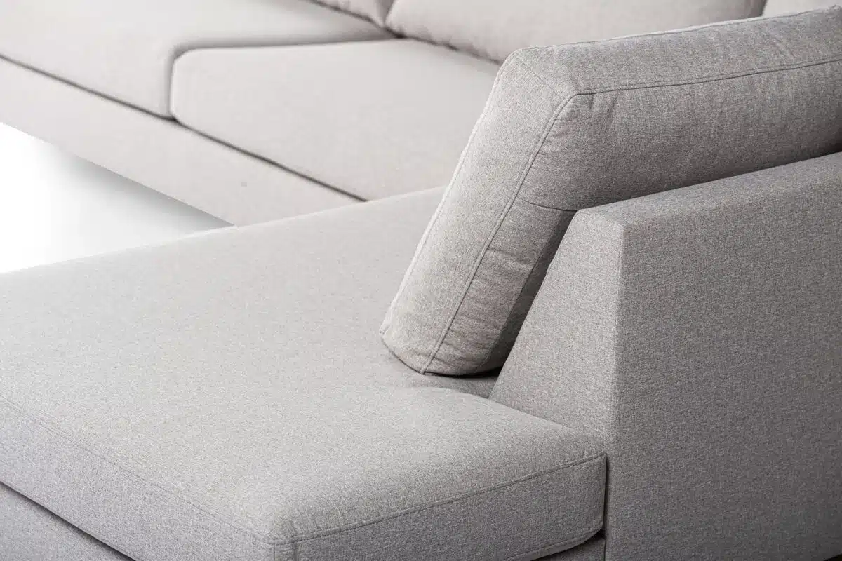 Close-up van een moderne grijze stoffen bank met een hoekige rugleuning en zachte kussens. Het sectionele stuk heeft strakke lijnen en een minimalistisch design, wat de eigentijdse stijl en het comfort benadrukt.