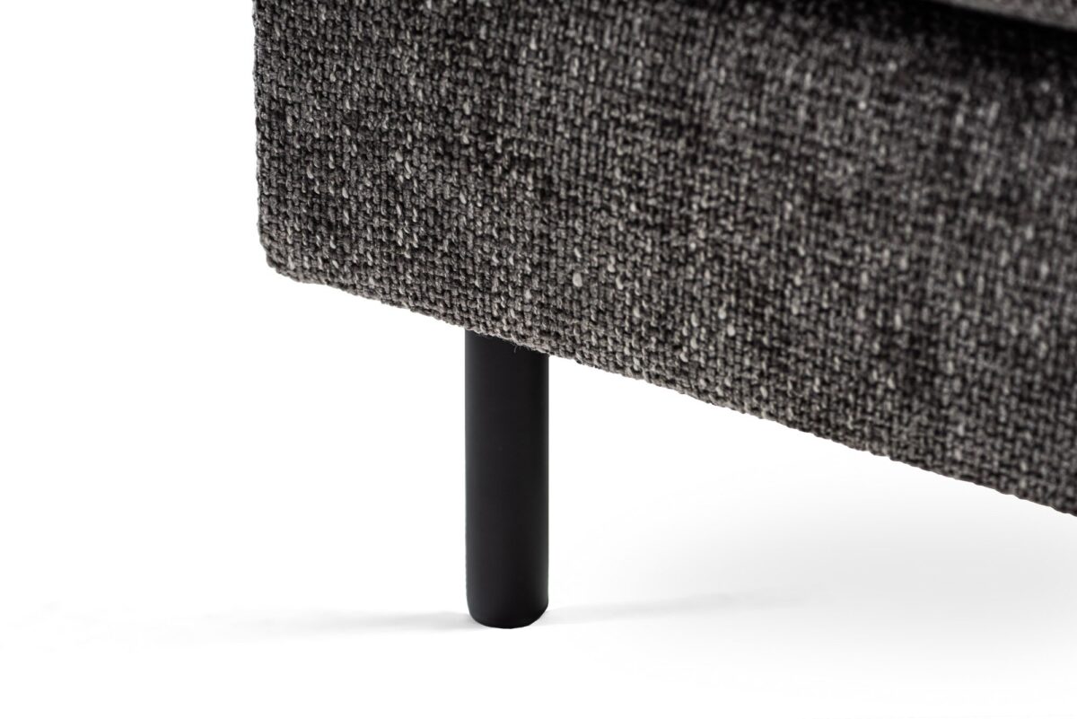Close-up van een donkergrijze getextureerde hoekbank met een zwarte cilindrische poot, tegen een witte achtergrond. De bekleding heeft een grove stoffen uitstraling.
