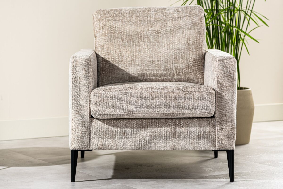Een beige gestoffeerde fauteuil met een structuurstof staat op een lichtgrijze betegelde vloer. Het heeft rechte armleuningen en zwarte poten. Een groene plant in een beige pot is zichtbaar op de achtergrond.