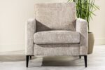 Een beige gestoffeerde fauteuil met een structuurstof staat op een lichtgrijze betegelde vloer. Het heeft rechte armleuningen en zwarte poten. Een groene plant in een beige pot is zichtbaar op de achtergrond.