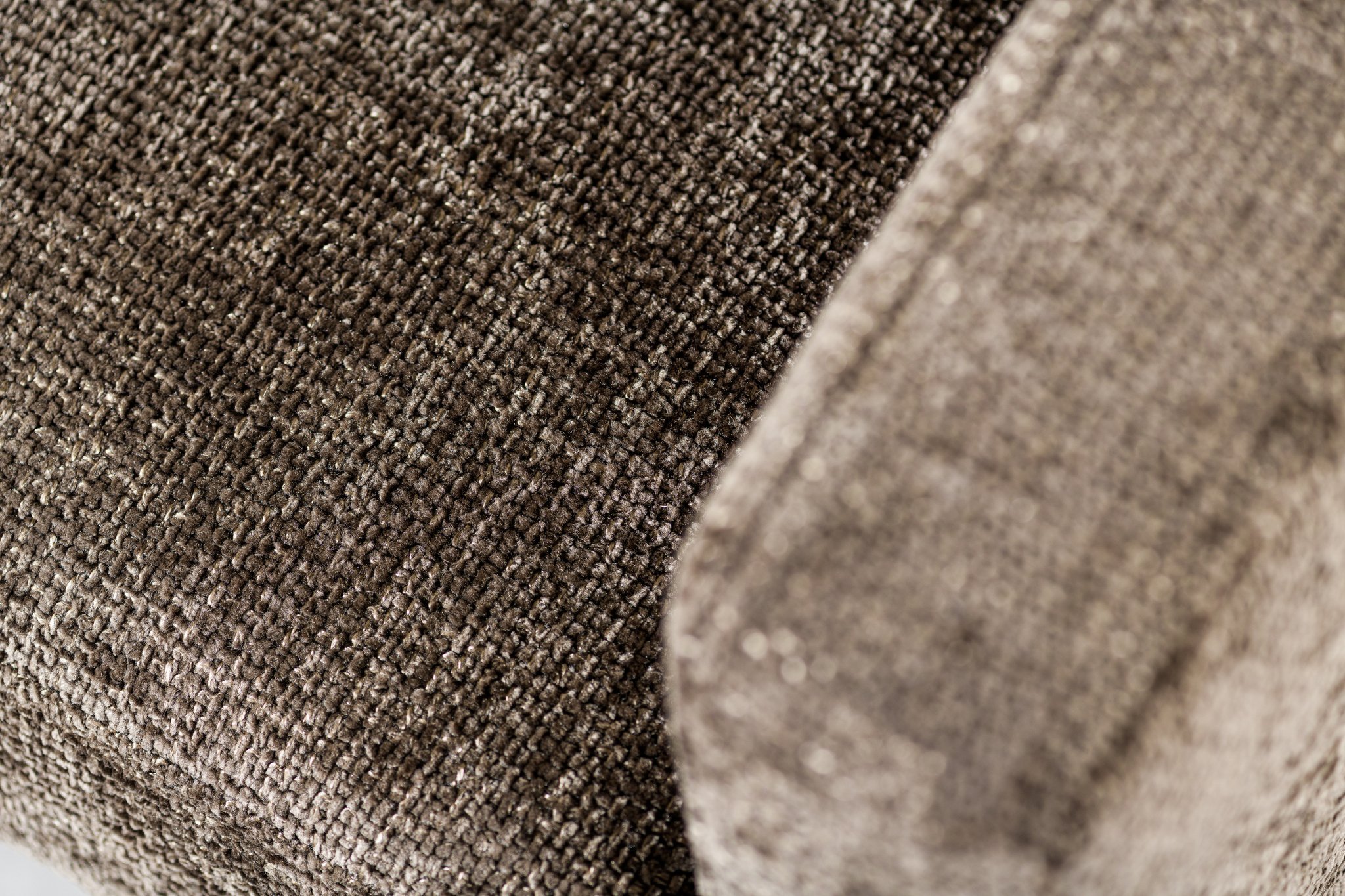 Close-up van een getextureerde stoffen bekleding, waarschijnlijk onderdeel van een armleuning van een bank of stoel, in tinten bruin en beige. De weving van de stof creëert een subtiel patroon dat het oppervlak een zachte, pluche uitstraling geeft.