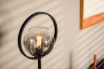 Een moderne lamp met een gloeiende bol in een heldere glazen bol, omgeven door een zwarte metalen ring. De achtergrond heeft zachte, schaduwrijke strepen op een lichtgekleurde muur, gedeeltelijk met een ingelijst kunstwerk.