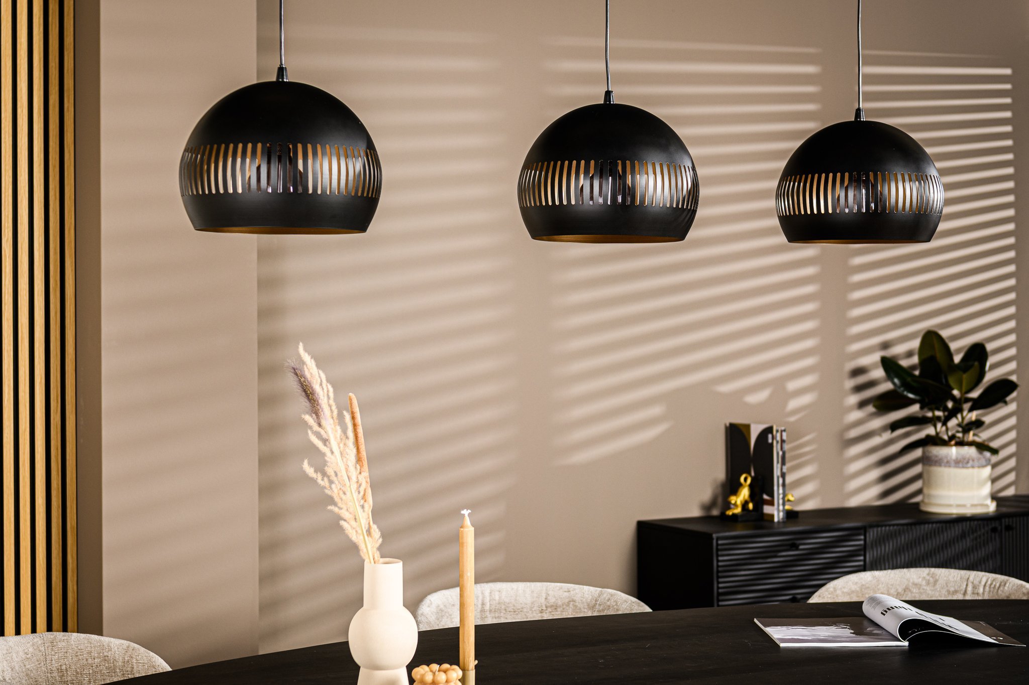 Moderne eetkamer met drie zwarte hanglampen die gestreepte schaduwen werpen op een beige muur. Een ronde tafel is gedekt met een vaas met droog gras, een kaars en een open tijdschrift. Een dressoir met decoratie-items en een plant staat op de achtergrond.