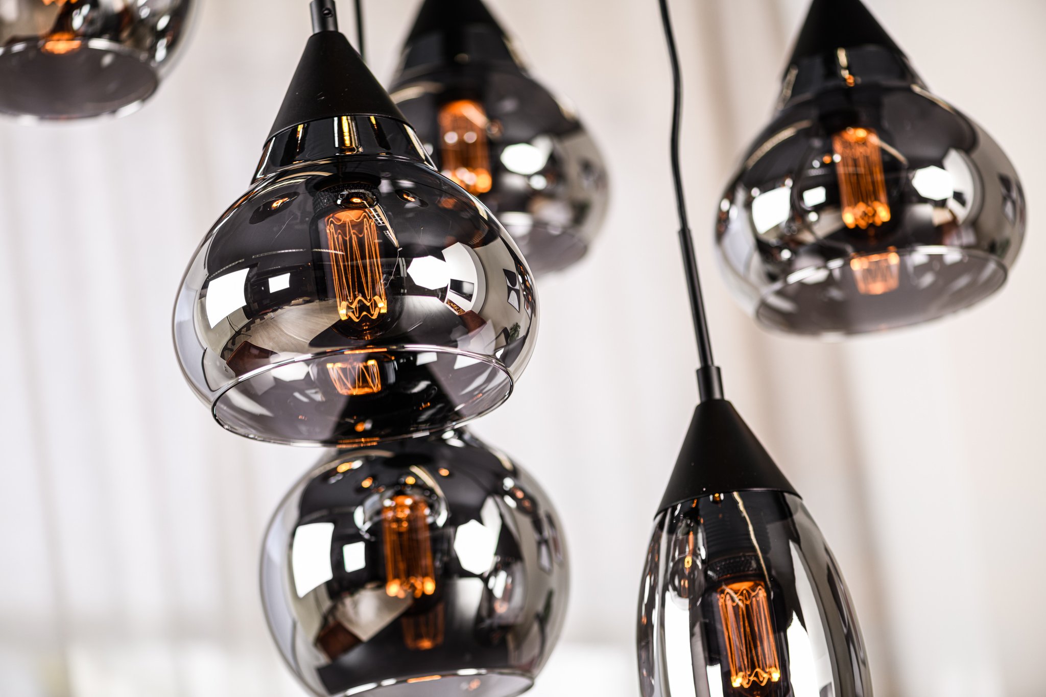 Close-up van meerdere moderne hanglampen met reflecterende chromen kappen en gloeiende gloeilampen, hangend aan zwarte snoeren. De achtergrond is wazig, met witte verticale lijnen die op gordijnen lijken.