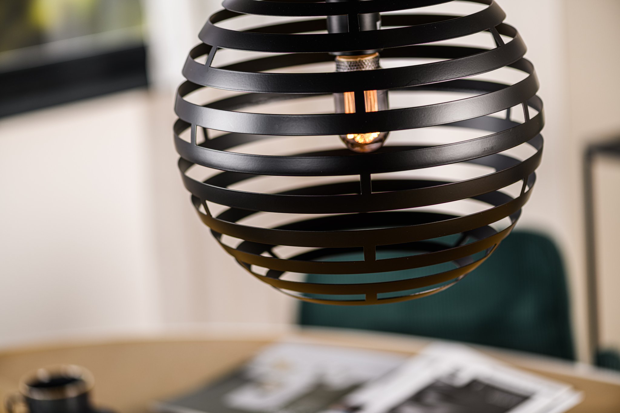 Close-up van een bolvormige hanglamp met zwarte horizontale strepen die een zichtbare lamp omsluiten. Hieronder is een wazige scène te zien met een deel van een tafel met een tijdschrift en een kopje, wat een moderne interieursetting suggereert.