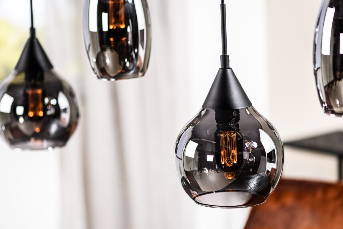 Close-up van stijlvolle hanglampen met zwart getinte glazen kappen en vintage-stijl filamentlampen. Ze hangen op verschillende hoogtes en werpen een warme gloed. Een wazige achtergrond van witte gordijnen en houten oppervlakken is zichtbaar.