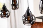 Close-up van stijlvolle hanglampen met zwart getinte glazen kappen en vintage-stijl filamentlampen. Ze hangen op verschillende hoogtes en werpen een warme gloed. Een wazige achtergrond van witte gordijnen en houten oppervlakken is zichtbaar.
