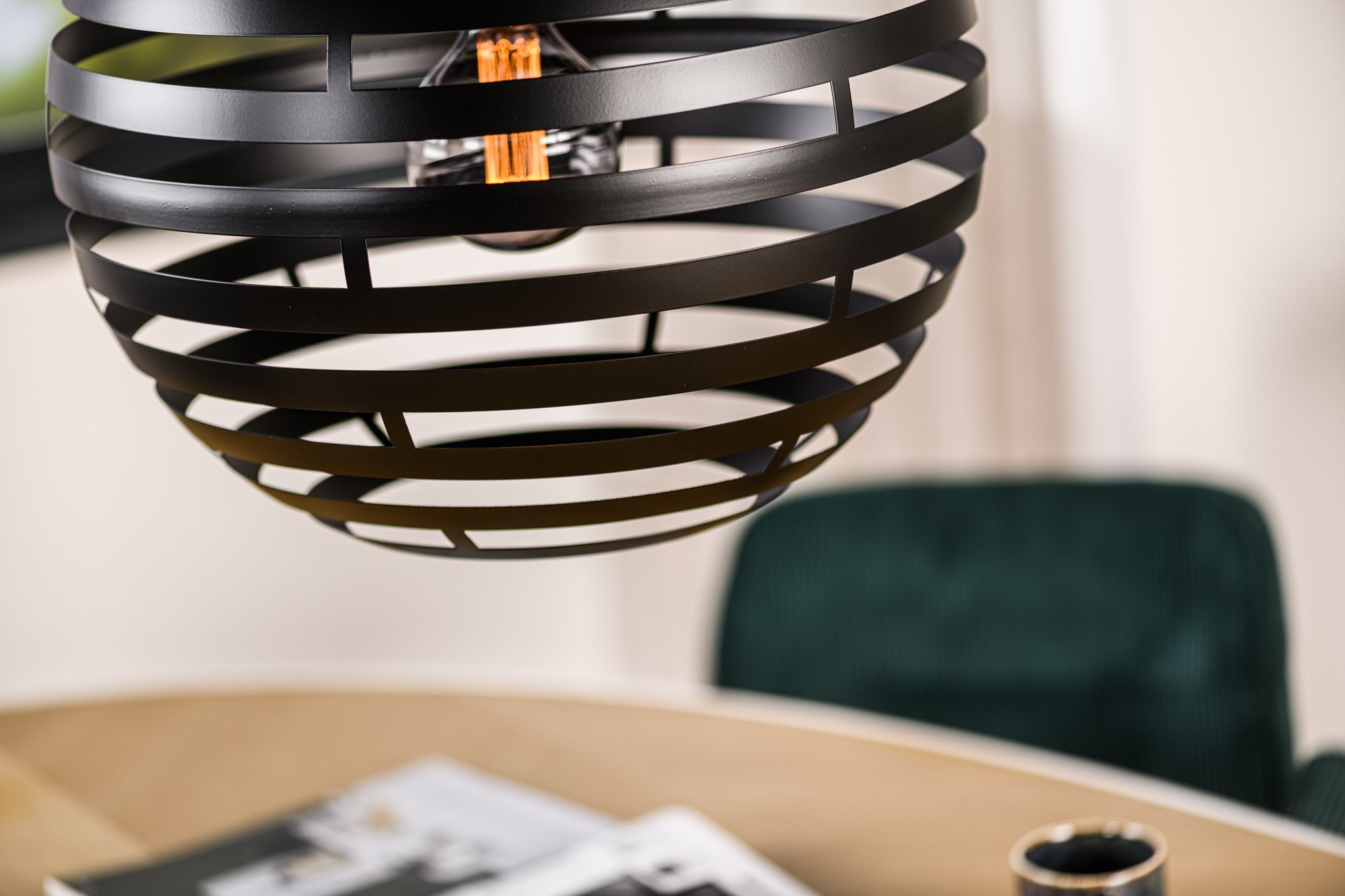 Close-up van een moderne zwarte bolvormige hanglamp met horizontale lamellen die boven een houten tafel hangt. Een donkergroene stoel en een opengeslagen tijdschrift zijn zichtbaar op de wazige achtergrond, wat een stijlvolle interieursetting suggereert.