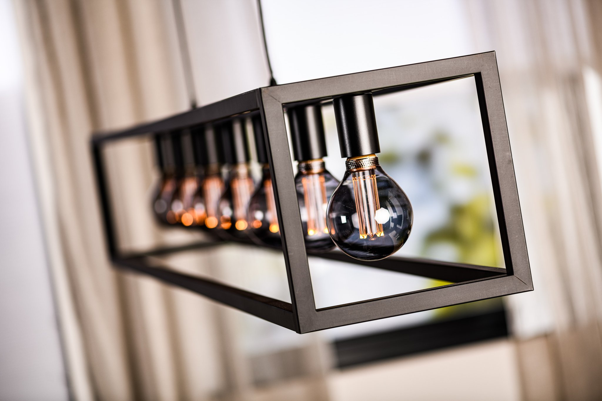 Close-up van een moderne rechthoekige hanglamp met zes hangende lampen, die elk warm gloeien. De lamp heeft een zwart metalen frame en is binnen geplaatst, met wazige gordijnen en een raam op de achtergrond.