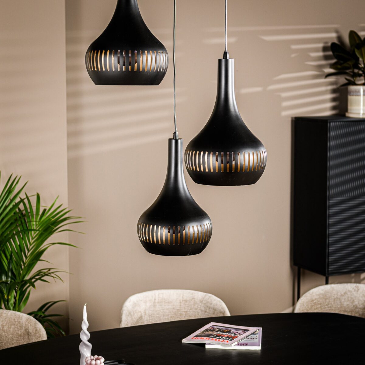 Drie moderne zwarte hanglampen hangen boven een eettafel met een donker oppervlak. Er ligt een tijdschrift op de tafel, met pluche beige stoelen eromheen. Een potplant en kast staan op de achtergrond, met schaduwen op de beige muur.