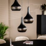 Drie moderne zwarte hanglampen hangen boven een eettafel met een donker oppervlak. Er ligt een tijdschrift op de tafel, met pluche beige stoelen eromheen. Een potplant en kast staan op de achtergrond, met schaduwen op de beige muur.