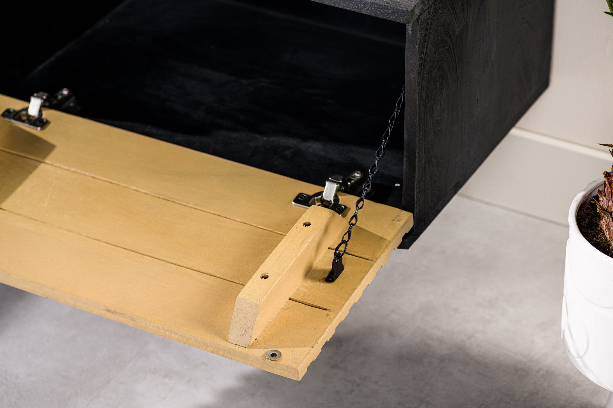 Een close-up van een houten klaptafel die gedeeltelijk is geopend, met metalen scharnieren en een ondersteunende ketting. De tafel heeft een lichtbruine kleur, die contrasteert met het donkere interieur van een kast. Rechts is een potplant zichtbaar.