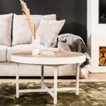 De Lennox Salontafel rond 74 cm, L350 eiken met een licht houten blad en witte poten staat op een groen vloerkleed voor een beige bank. Op tafel staan een witte vaas met gedroogd gras, een groene kaars en een boek.