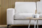 De 3 zits bank CL L+R, stof Valente, V920 naturel, een moderne lichtbeige bank, staat voor een houten lattenwand met een ronde bijzettafel ernaast met een witte vaas, kaars en decoratieve voorwerpen.
