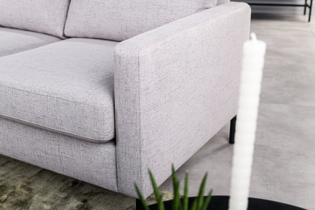 Close-up van de 3 zits bank CL L+R in Valente V311 grijs stof met vierkante armleuning, getoond met een groen vloerkleed, plant en witte structuurkaars op een zwarte ronde tafel. De setting heeft een lichtgekleurde tegelvloer.