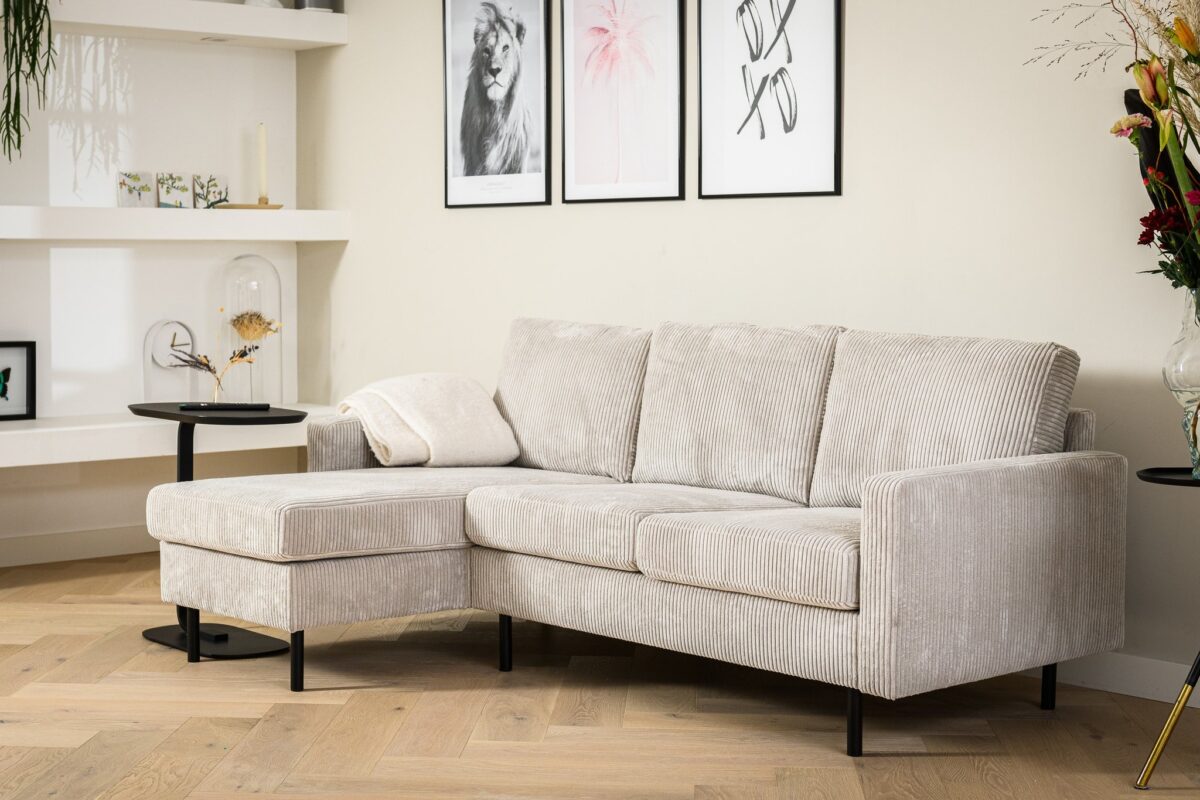 Een moderne woonkamer heeft een lichtgrijze hoekbank met een chaise longue. De ruimte omvat twee zwarte bijzettafels en ingelijste kunstwerken aan de muur. Decoratieve items en planten sieren open planken en de vloer is van hout.
