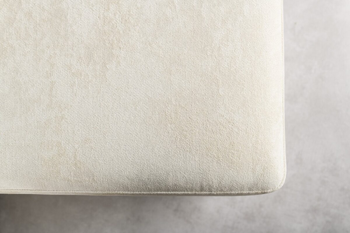 Close-up van een beige stoffen hoek van een kussen of gestoffeerd meubelstuk tegen een lichtgrijze achtergrond. De textuur lijkt zacht en licht getextureerd.