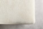 Close-up van een beige stoffen hoek van een kussen of gestoffeerd meubelstuk tegen een lichtgrijze achtergrond. De textuur lijkt zacht en licht getextureerd.