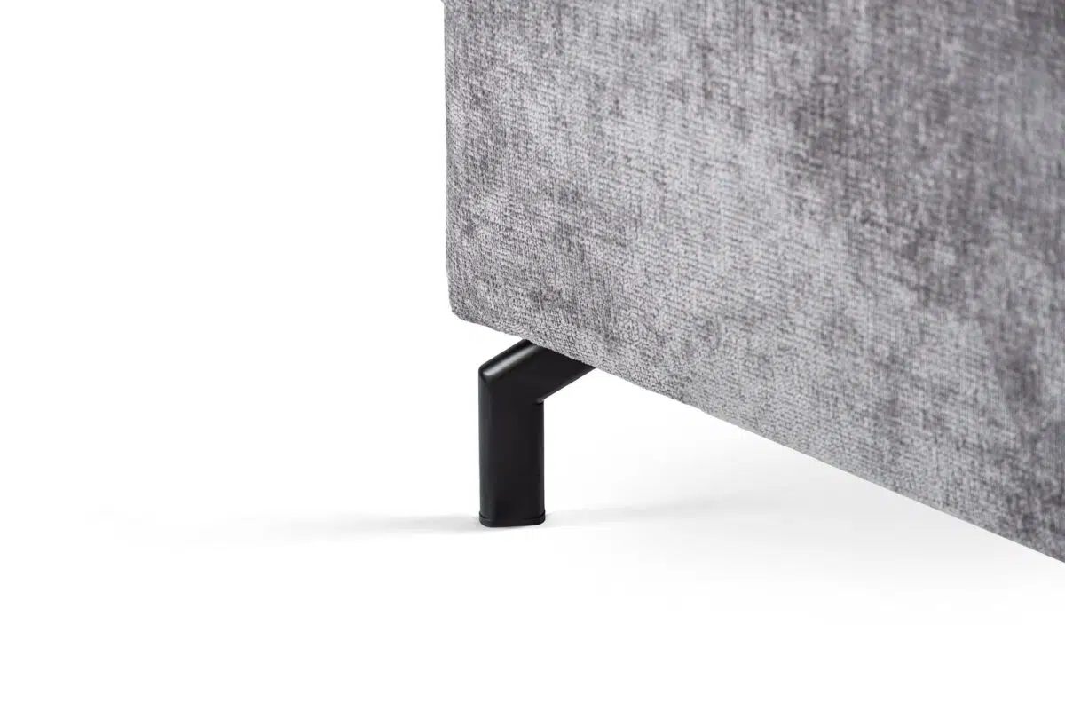 Close-up van een grijs stoffen meubel met een metalen poot. De textuur van de stof is zichtbaar zacht en licht glanzend. De metalen poot is zwart en heeft een modern, hoekig design. De achtergrond is effen wit.