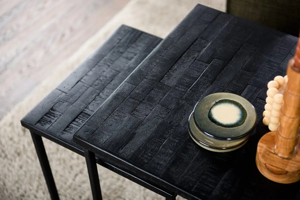 Twee zwarte vierkante tafels met een gestructureerd oppervlak staan naast elkaar. Op één tafel liggen keramische borden met een groenige tint op elkaar. Een deel van een houten decoratief item is rechts zichtbaar. Er ligt een beige tapijt onder.