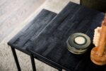 Twee zwarte vierkante tafels met een gestructureerd oppervlak staan naast elkaar. Op één tafel liggen keramische borden met een groenige tint op elkaar. Een deel van een houten decoratief item is rechts zichtbaar. Er ligt een beige tapijt onder.