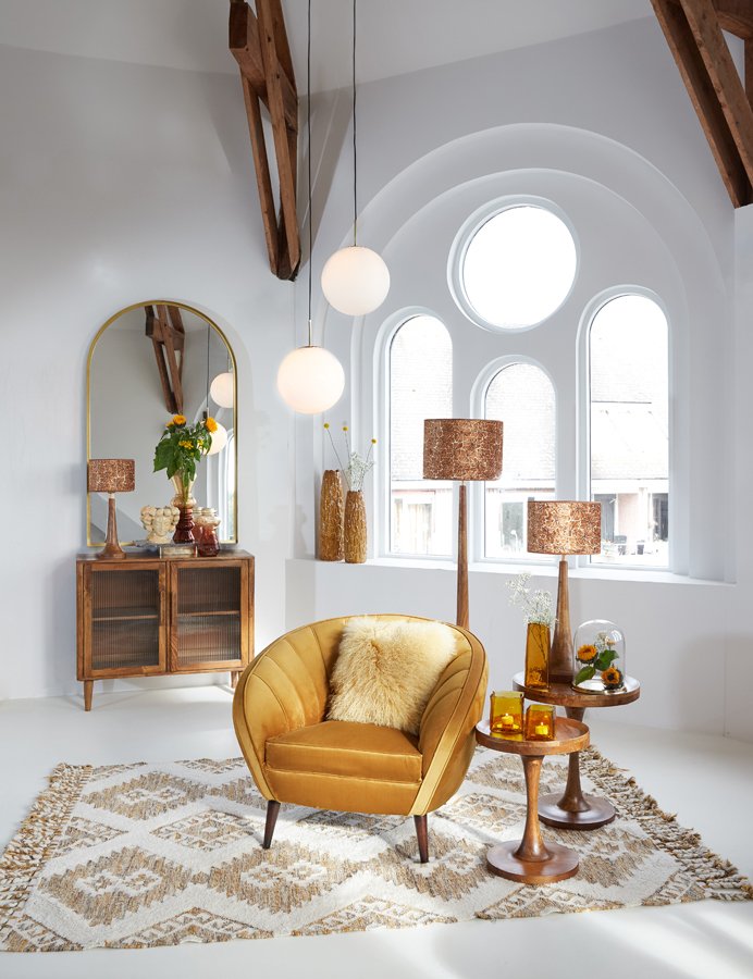 Een gezellige kamer met een mosterdgele stoel, een geometrisch kleed en een ronde bijzettafel met lampen. Een groot boograam en spiegel brengen licht in de ruimte, versierd met vazen en planten voor een mid-century moderne sfeer.