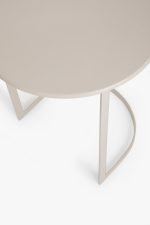 Een ronde beige tafel met een minimalistisch design heeft een unieke, gebogen pootondersteuning. Het tafelblad heeft een gladde afwerking en de achtergrond is een eenvoudig, lichtgrijs, wat de moderne stijl van de tafel benadrukt.