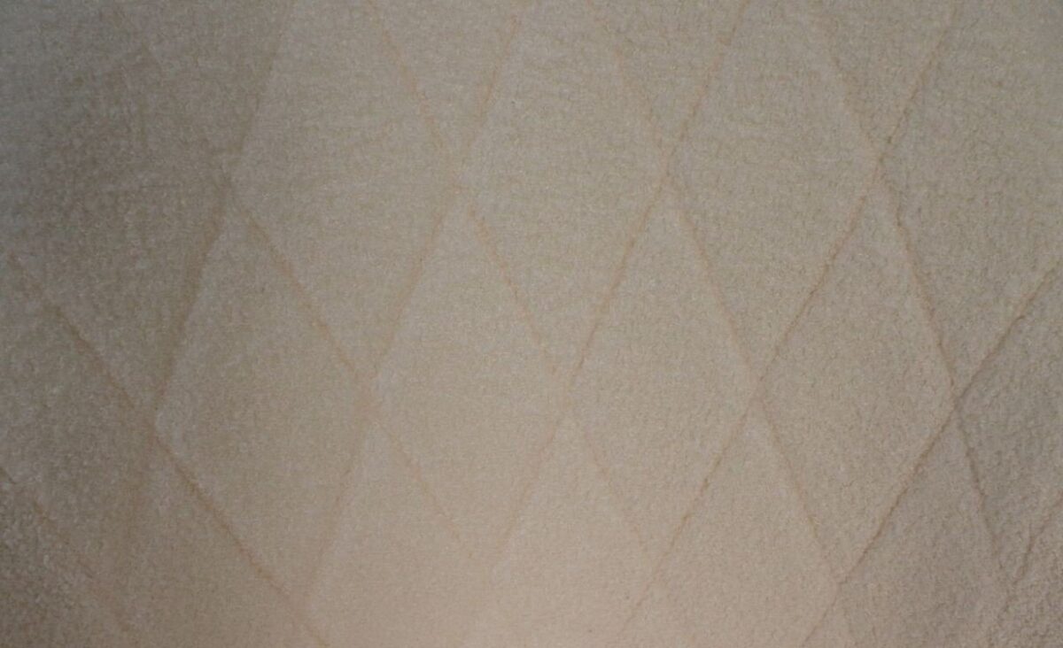 Een close-up van een zachte beige stof met een subtiel ruitpatroon. De textuur lijkt pluche, en lijkt op een warme deken of stoffering.