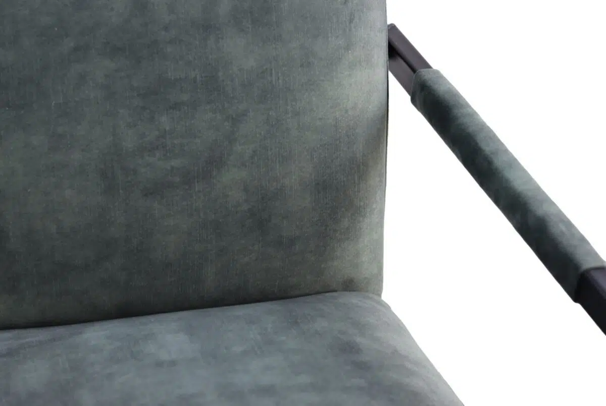 Close-up van een stoel met een donkergroene, getextureerde stof en zwarte metalen armleuningen. De focus ligt op de rugleuning en een deel van de zitting, tegen een effen witte achtergrond.