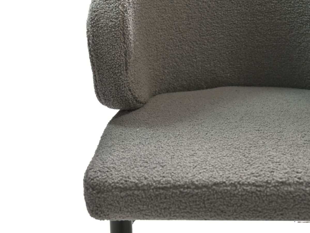 Close-up van een grijze, getextureerde fauteuil met een gebogen rugleuning. De stof lijkt zacht en heeft een bouclé-textuur. De stoel heeft metalen poten, gedeeltelijk zichtbaar aan de onderkant.