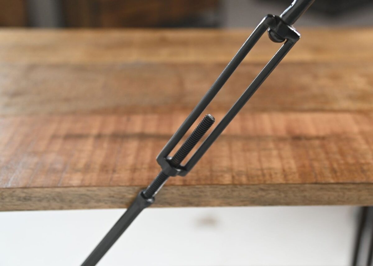 Close-up van een metalen spanstang die een houten oppervlak vastklemt, met de focus op het schroefmechanisme met schroefdraad. De achtergrond toont de houtstructuur en de rand van de tafel.
