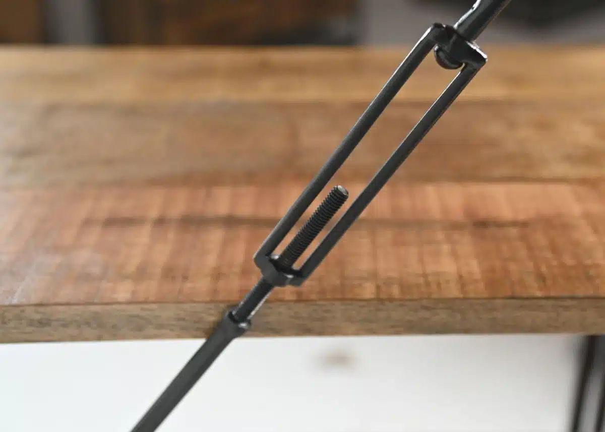 Close-up van een metalen spanstang die een houten oppervlak vastklemt, met de focus op het schroefmechanisme met schroefdraad. De achtergrond toont de houtstructuur en de rand van de tafel.