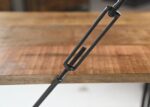 Close-up van een metalen spanstang die een houten oppervlak vastklemt, met de focus op het schroefmechanisme met schroefdraad. De achtergrond toont de houtstructuur en de rand van de tafel.
