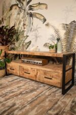 Een rustieke houten tv-standaard met lades en een zwart metalen frame staat op een gepatternde houten vloer. Een vintage radio staat op de middelste plank. Potplanten staan erop en eromheen, met een groot bladbehang op de achtergrond.