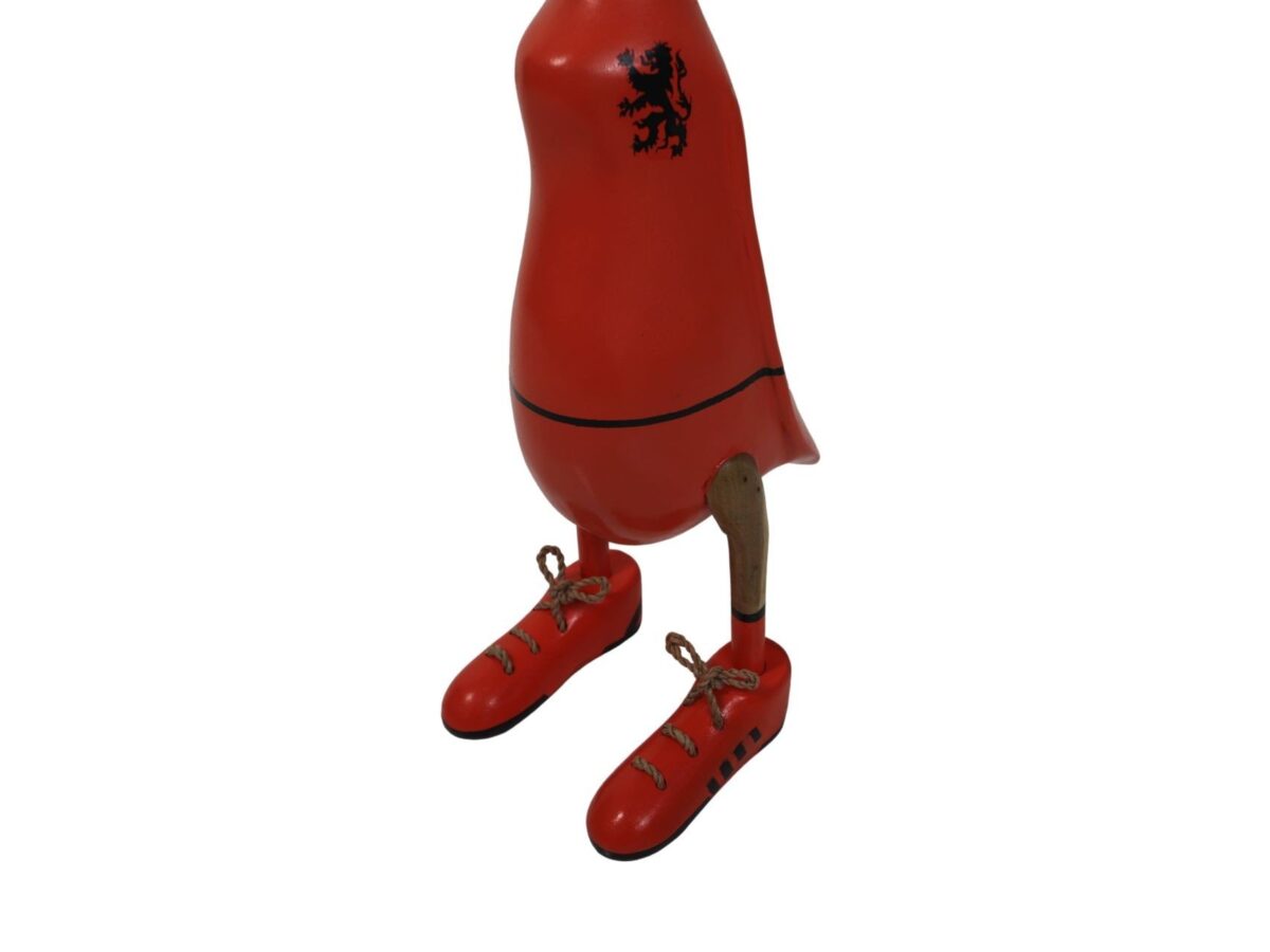 Een rood houten beeld dat lijkt op een vogel met een zwarte leeuwenkam op zijn borst. Het heeft houten poten en rode schoenen met veters. Het bovenste deel van het beeld heeft geen kop of zichtbare vleugels.