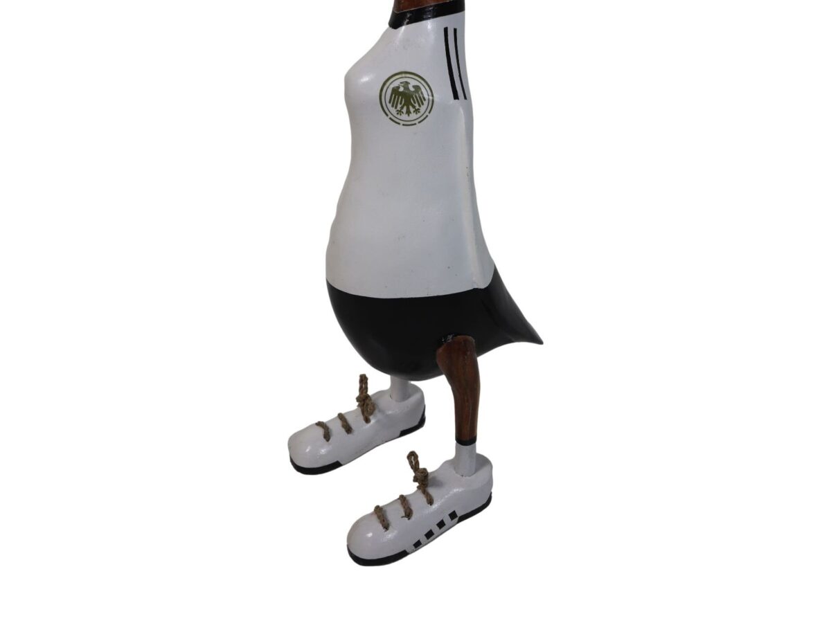 Een houten eendfiguur gekleed in een wit en zwart sporttenue met een wapen, dat lijkt op een voetbalteamuniform. De eend draagt witte houten sneakers met zwarte strepen en gouden noppen.